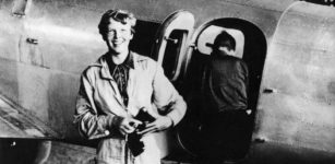 American aviator/pilot Amelia Earhart (1897-1937) standing by her Lockheed Electra dressed in overalls, with Fred Noonan getting into the plane in the background.